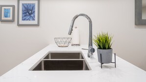 A modern kitchen with a stainless steel sink, a white marble countertop, a wire basket, a roll of paper towels, and a potted plant.