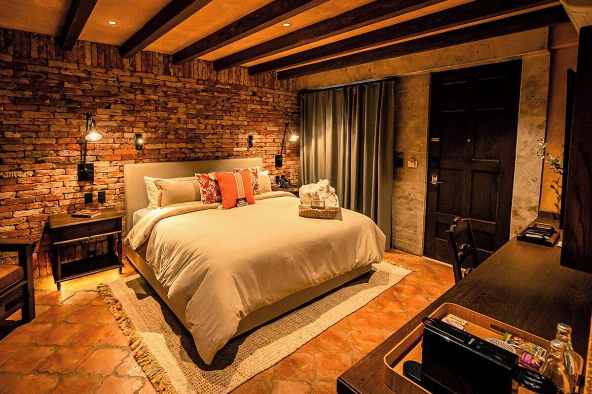 A cozy bedroom with a brick wall, a large bed with pillows, bedside lamps, and a wooden nightstand.