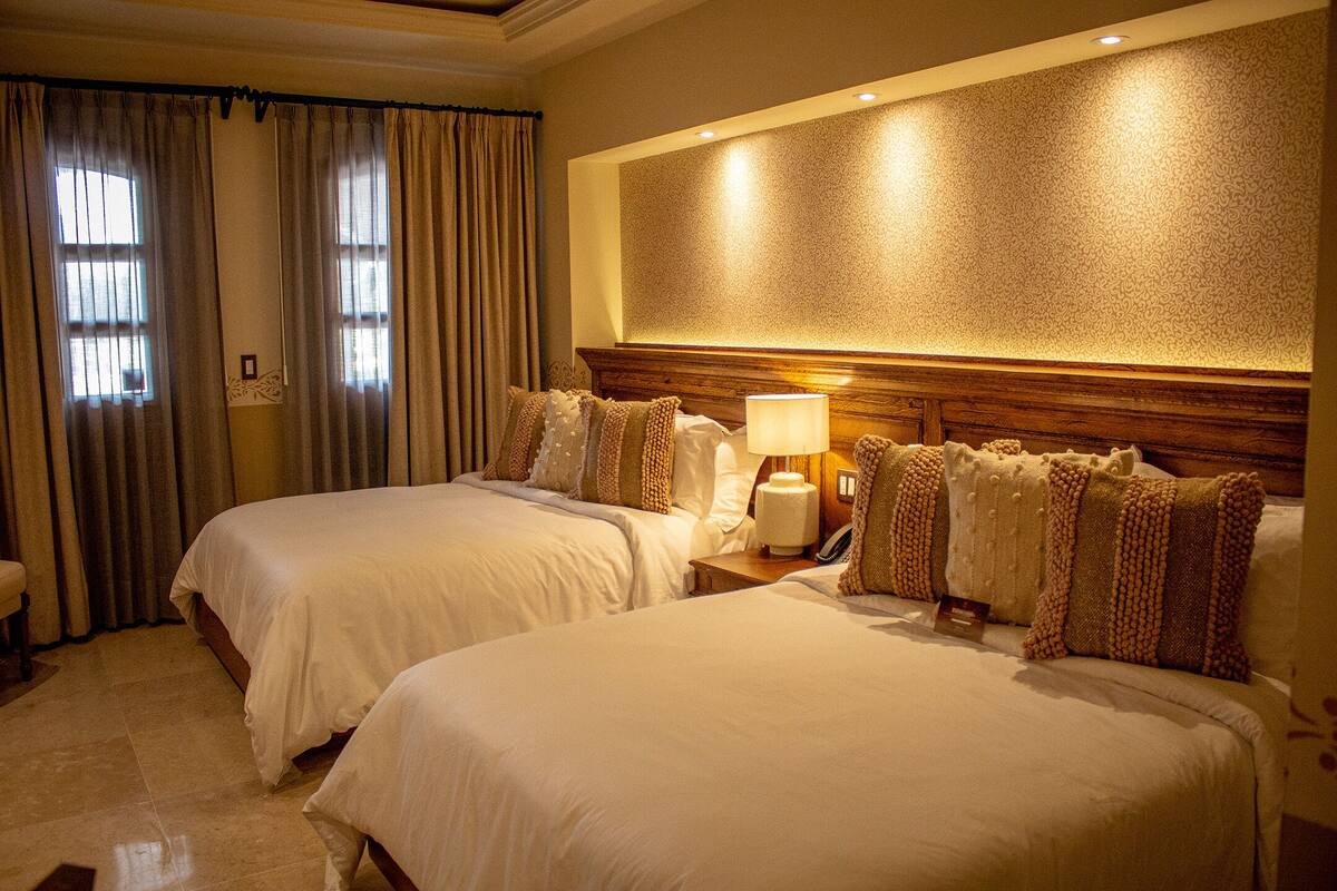 A hotel room with two beds, a headboard with a textured wall, a bedside lamp, and a window with curtains.