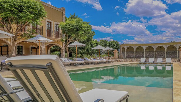 Zona de piscina con sillas de descanso y sombrillas, rodeada por un edificio con arcos y balcones.