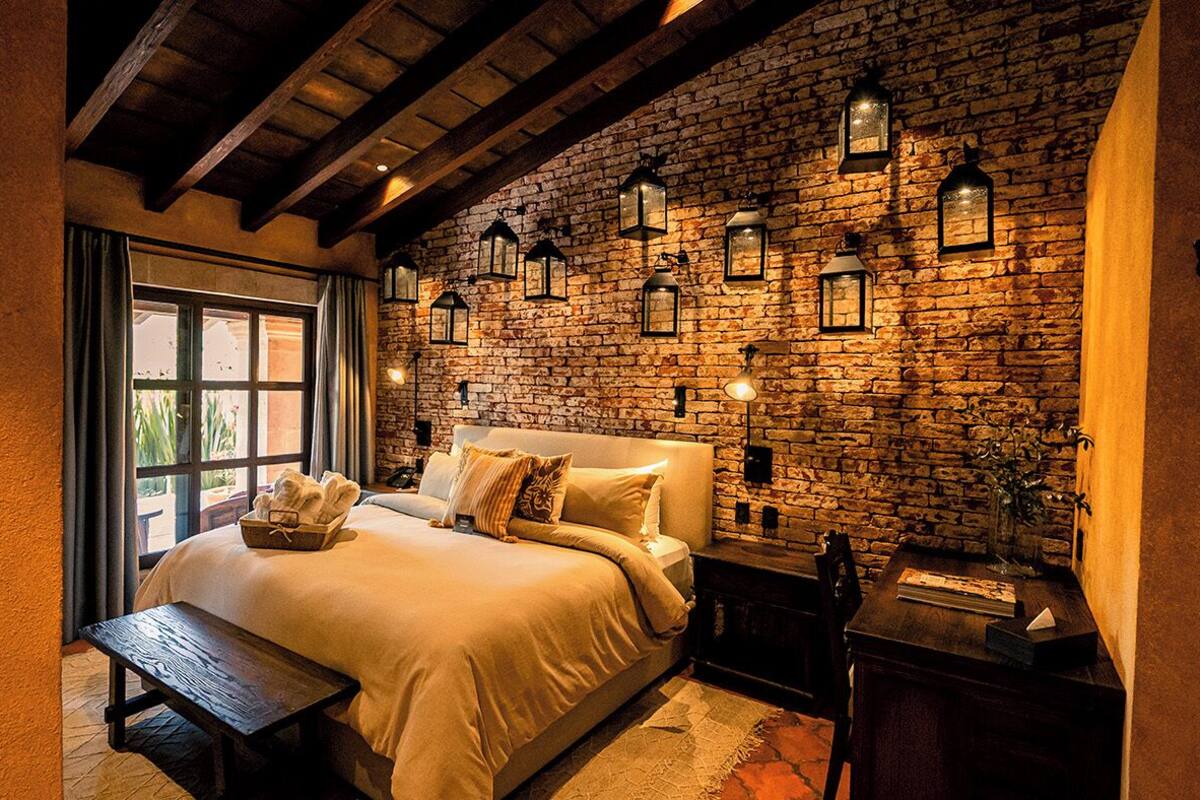 A cozy bedroom with a stone wall, a large bed, a wooden bench, and wall-mounted lanterns.