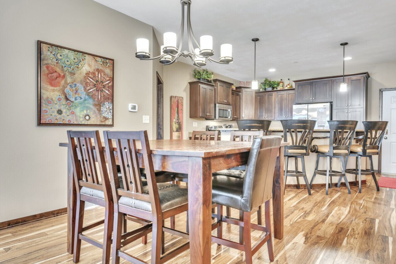 A dining area with a wooden table and chairs, a chandelier, and a kitchen in the background.