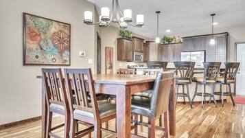 A dining area with a wooden table and chairs, a chandelier, and a kitchen in the background.