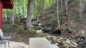 Eine rustikale Holzbank und ein Picknicktisch in einer bewaldeten Gegend mit einem kleinen Bach und einem roten Gebäudebau.