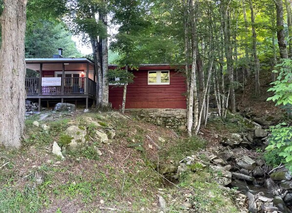 Una cabaña rústica con terraza de madera y un ventanal pequeño, rodeada de un bosque denso y un arroyo pedregoso.