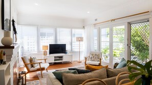 A modern living room with a flat-screen TV, comfortable seating, and large windows. - Yanderra - Discover relaxation at this charming cottage opposite the beachside golf course (Mollymook)