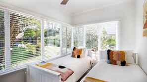 A bedroom with two beds, white bedding, and a view of greenery through large windows. - Yanderra - Discover relaxation at this charming cottage opposite the beachside golf course (Mollymook)