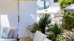 A patio area with a white wall, a cushioned seating area, and a potted plant. - Yanderra - Discover relaxation at this charming cottage opposite the beachside golf course (Mollymook)