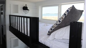 Une chambre à coucher avec un lit, une fenêtre donnant sur la plage, une rambarde noire et un luminaire fixé au mur.