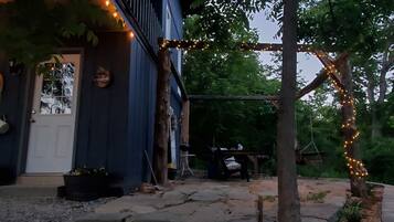 A rustic outdoor area with a stone pathway leading to a wooden door, surrounded by greenery and string lights.