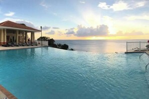 Een zwembad met uitzicht op de oceaan bij zonsondergang, een overdekt terras met loungebedden en een omheind terras.