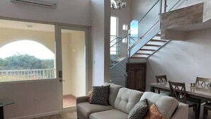 Ein geräumiger Wohnbereich mit grauem Sofa, einem Esstisch und einer Treppe, die in eine obere Ebene führt.