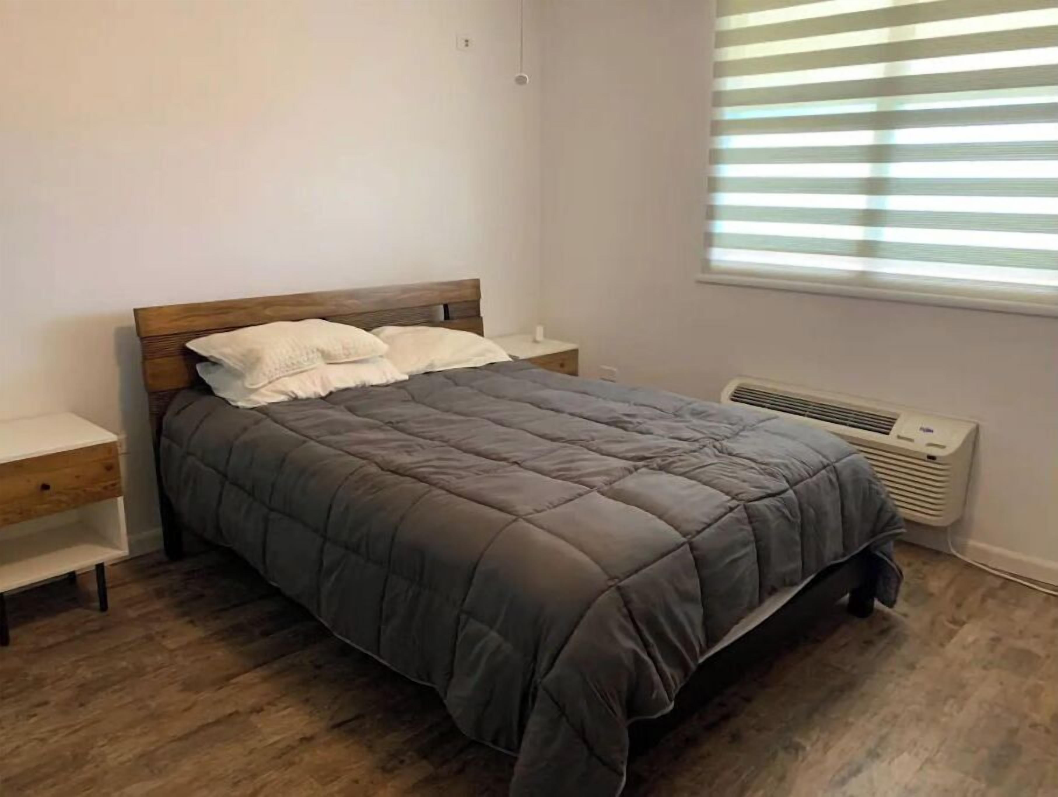 A bedroom with a bed, wooden nightstands, a window with blinds, and a wall-mounted air conditioner.