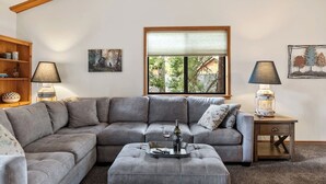 A cozy living room with a grey sectional sofa, a wooden side table with a lamp, and a window with a view of trees.
