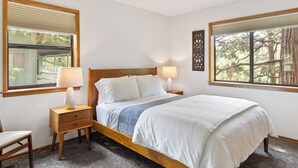 A bedroom with a wooden bed, two bedside tables with lamps, a chair, and windows with blinds.