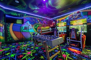A brightly lit arcade room with a foosball table, racing game machines, and a large screen displaying a video game. - Amazing New Villa!  Exceptionally Maintained! Fun for the Whole Family! (Four Corners)