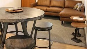 Un salon avec un canapé en cuir marron, une table de salle à manger ronde entourée de tabourets et, au fond, une porte en bois.
