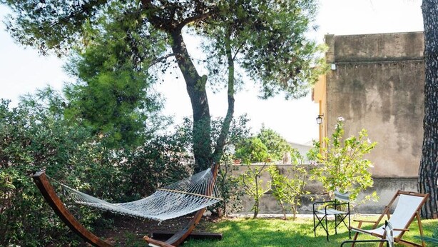 A hammock, two wooden chairs, and a small table in a garden with trees and a concrete wall in the background. - Casa Guardia Bilocal Apartment Near the sea (Acireale)
