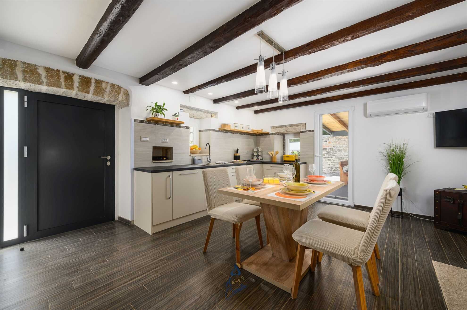 A modern kitchen with wooden beams, a dining area with a wooden table and chairs, and a stone wall visible through the door.