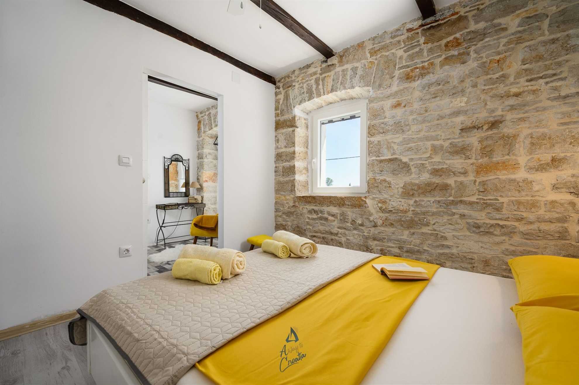 Une chambre avec un mur en pierre, un lit avec une literie jaune, une fenêtre et un miroir.