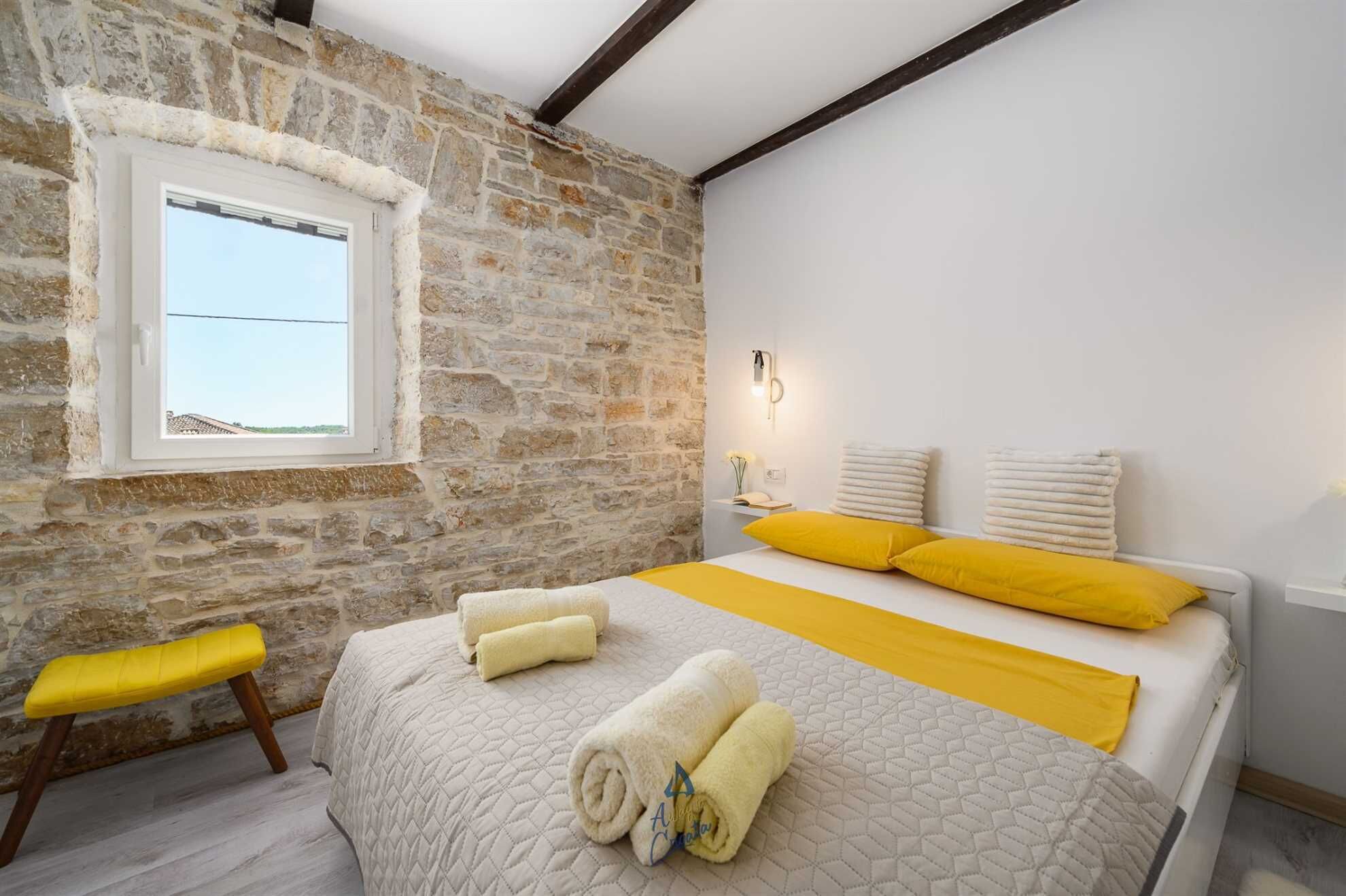 Un dormitorio con pared de piedra, cama con ropa de cama amarilla y blanca, un banco y una ventana.