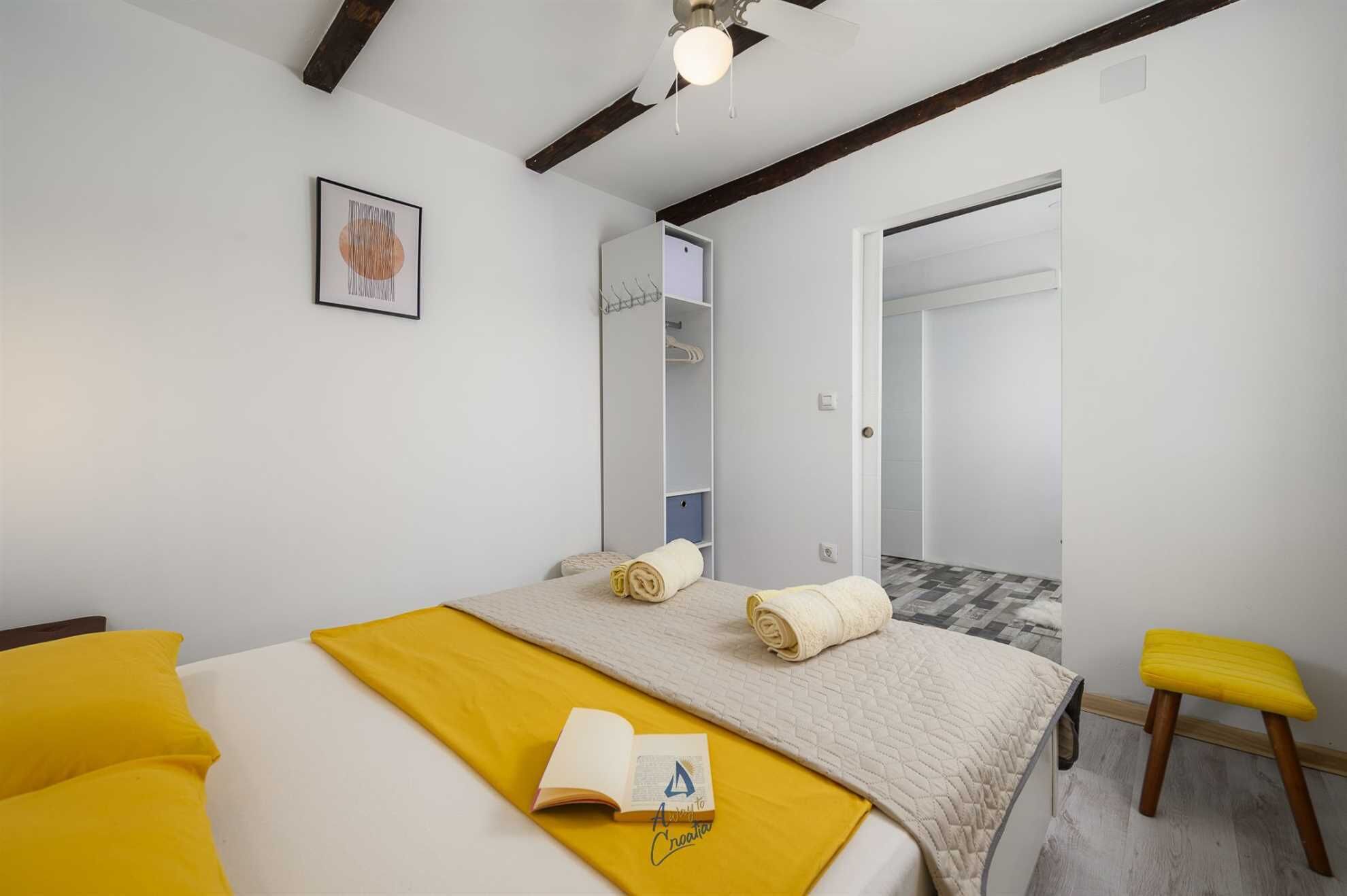 A bedroom with a bed, a bedside table with a book, a yellow chair, a wardrobe, and a door leading to another room.