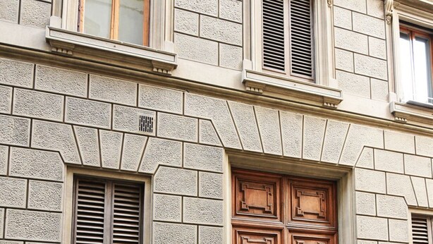 Une façade d’immeuble avec une grande porte à double battant en bois, des murs à l’aspect pierre et de nombreuses fenêtres dotées de volets.