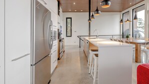 Et moderne køkken med stålemhætter, en central ø med barstole, hængende lamper og udsigt ud af vinduet.