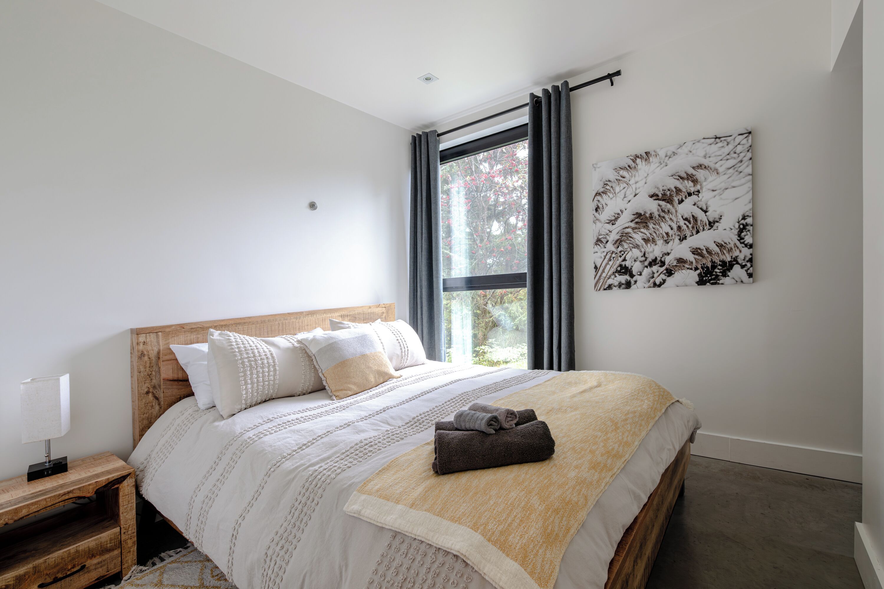 Ein Schlafzimmer mit einem hölzernen Bett, weißer Bettwäsche, einem Nachttisch, einem Fenster mit Vorhängen und einem an der Wand befestigten Kunstwerk.