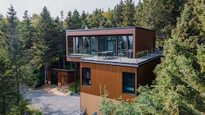 En moderne hytte med to etager, store vinduer, en terrasse og en omkringliggende skov.