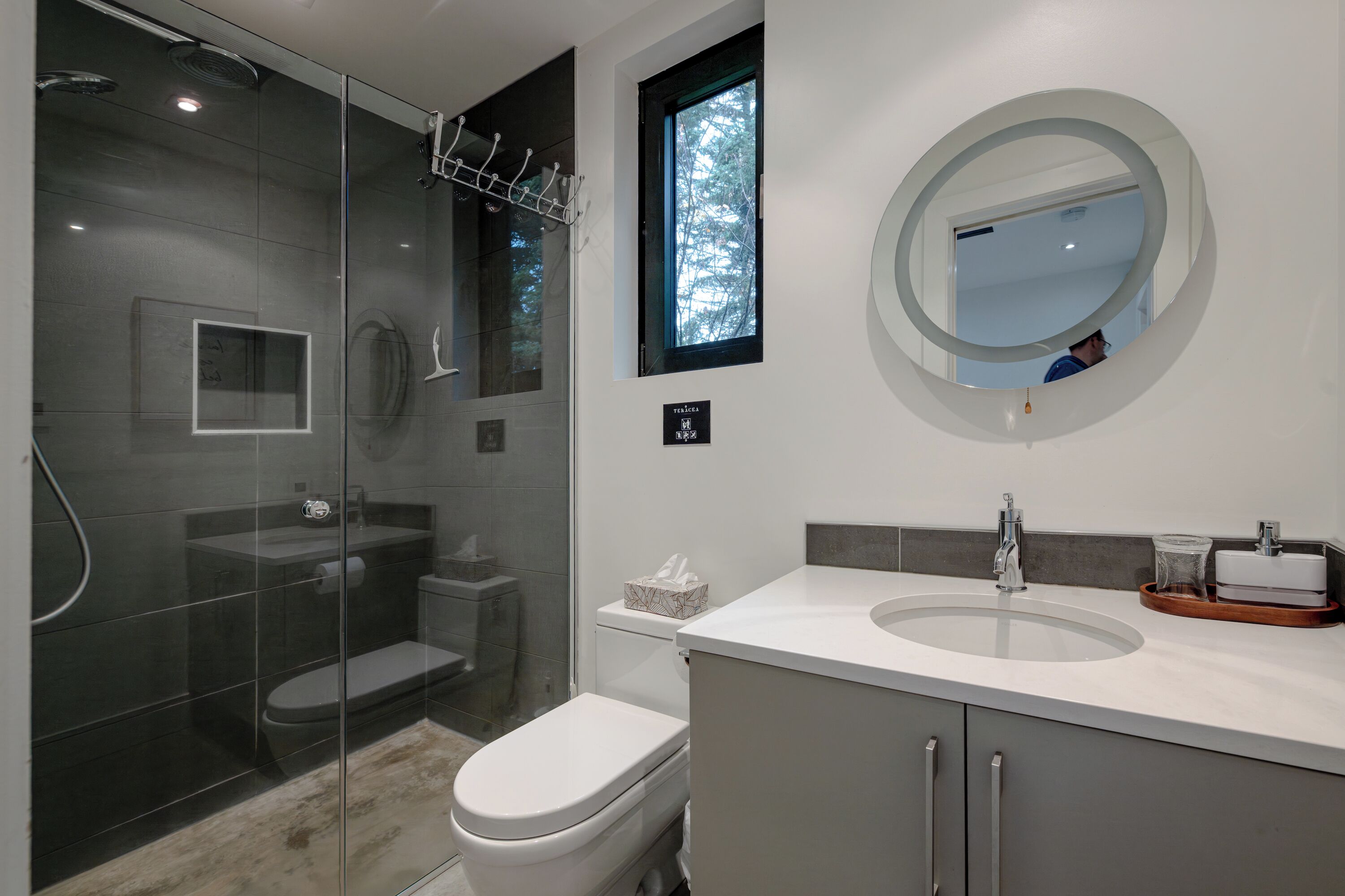 Ein modernes Badezimmer mit einer Glastrennwand-Dusche, einer Toilette, einem Waschbecken mit Spiegel und einem Fenster für natürliches Licht.