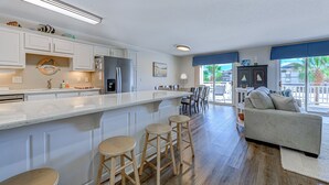 A modern kitchen with white cabinetry, a central island with stools, and a dining area with a gray sofa and chairs. - Recently Renovated Spacious 6 Bedroom Condo, Just Steps From The Ocean (Ocean Isle Beach)