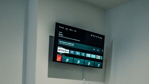 A modern living room with a mounted flat-screen TV displaying a streaming service interface. There is a decorative plant and a woven basket on the floor. - Bv Cosy Basement Apartment At Conditioning House (Bradford)