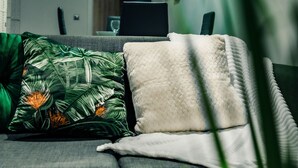 A modern living room with a grey sofa, a white throw blanket, and a green tropical-patterned pillow. A black column and a chandelier are visible in the background. - Bv Cosy Basement Apartment At Conditioning House (Bradford)