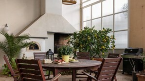 Een terras buiten met een ronde houten tafel, vier houten stoelen, een metselwerk haardvuur, potplanten en een barbecue.