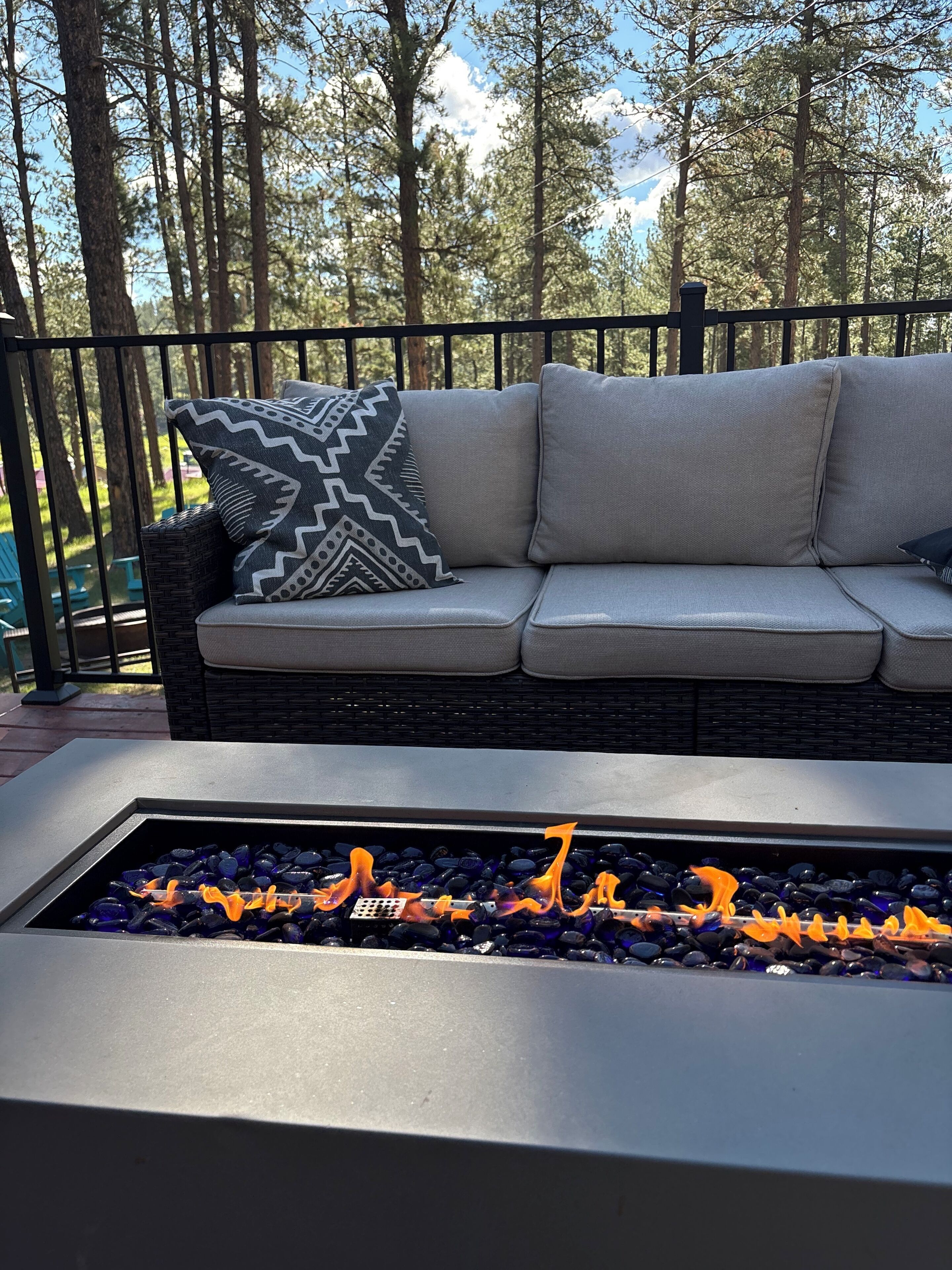 A fire pit with blue stones and orange flames, a grey outdoor sofa with patterned cushions, and a black metal railing.