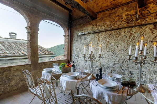 A dining area with a rustic stone wall, a table set for four, and a view of a tiled roof through an archway.