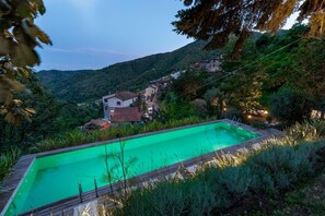 A rectangular swimming pool with a wooden deck, surrounded by lush greenery and overlooking a hillside village.