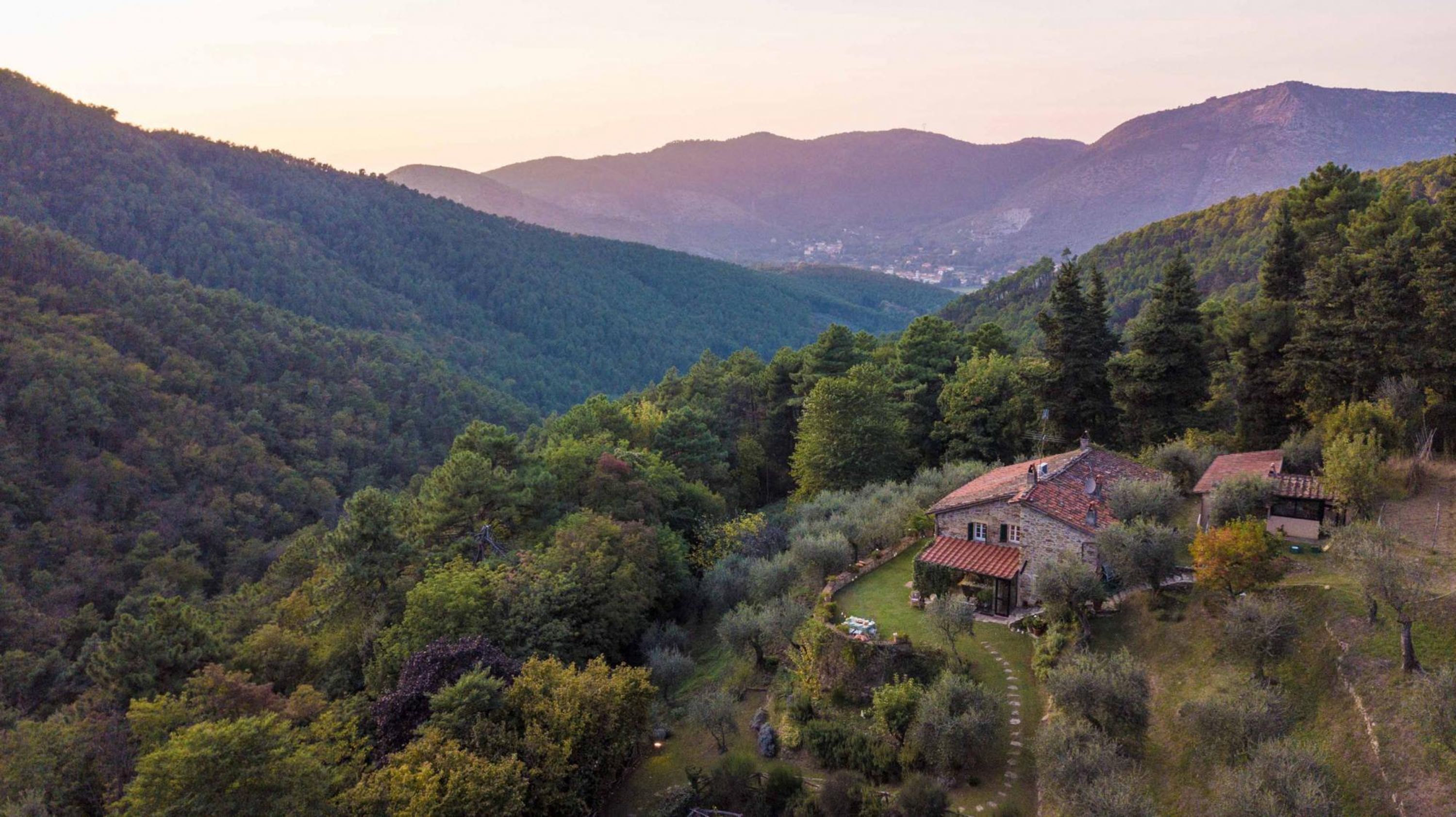 Una casa in pietra con tetto in tegole rosse, immersa in un giardino rigoglioso e una fitta foresta, con dolci colline sullo sfondo.