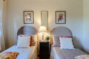 Una habitación con dos camas, cada una con cabecero, mesita de noche con lámpara y cuadros en la pared.