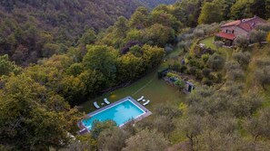 An aerial view of a property with a swimming pool surrounded by lush greenery, a stone house, and a well-maintained garden.