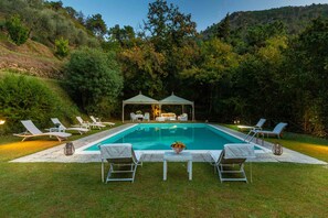 Una piscina rectangular con un pabellón en el centro, rodeada de sillones de descanso y una mesita baja con un frutero.