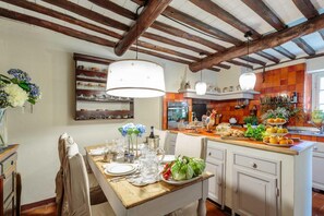 Una cocina con vigas de madera, una isla central con frutas y verduras, y una lámpara colgante.
