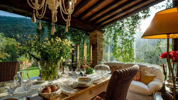 An outdoor dining area with a wooden table set for a meal, surrounded by wicker furniture and a view of a lush garden. - Casa Nel Bosco in Santa Maria del Giudice (Lucca)