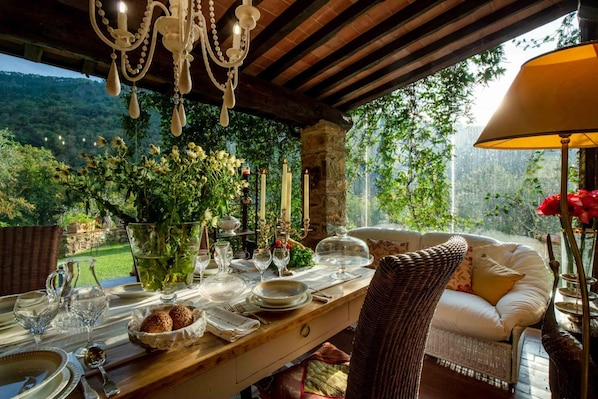 An outdoor dining area with a wooden table set for a meal, surrounded by wicker furniture and a view of a lush garden.