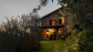 A stone house with a well-lit interior, surrounded by lush greenery and a stone pathway leading to the entrance. - Casa Nel Bosco in Santa Maria del Giudice (Lucca)