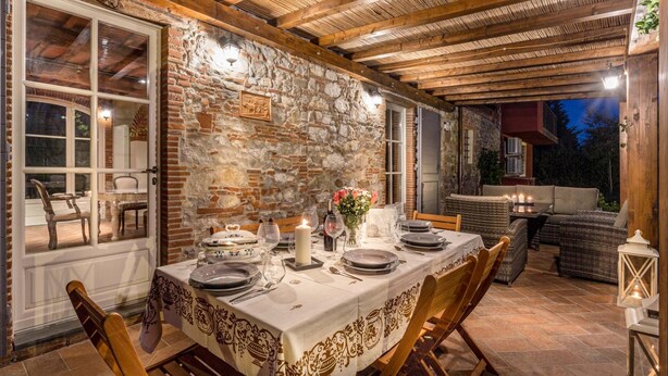 An outdoor dining area with a stone wall, wooden ceiling, and a table set for two with candles and a vase of flowers. - Villa sul Fiume in Monte San Quirico (Lucca)