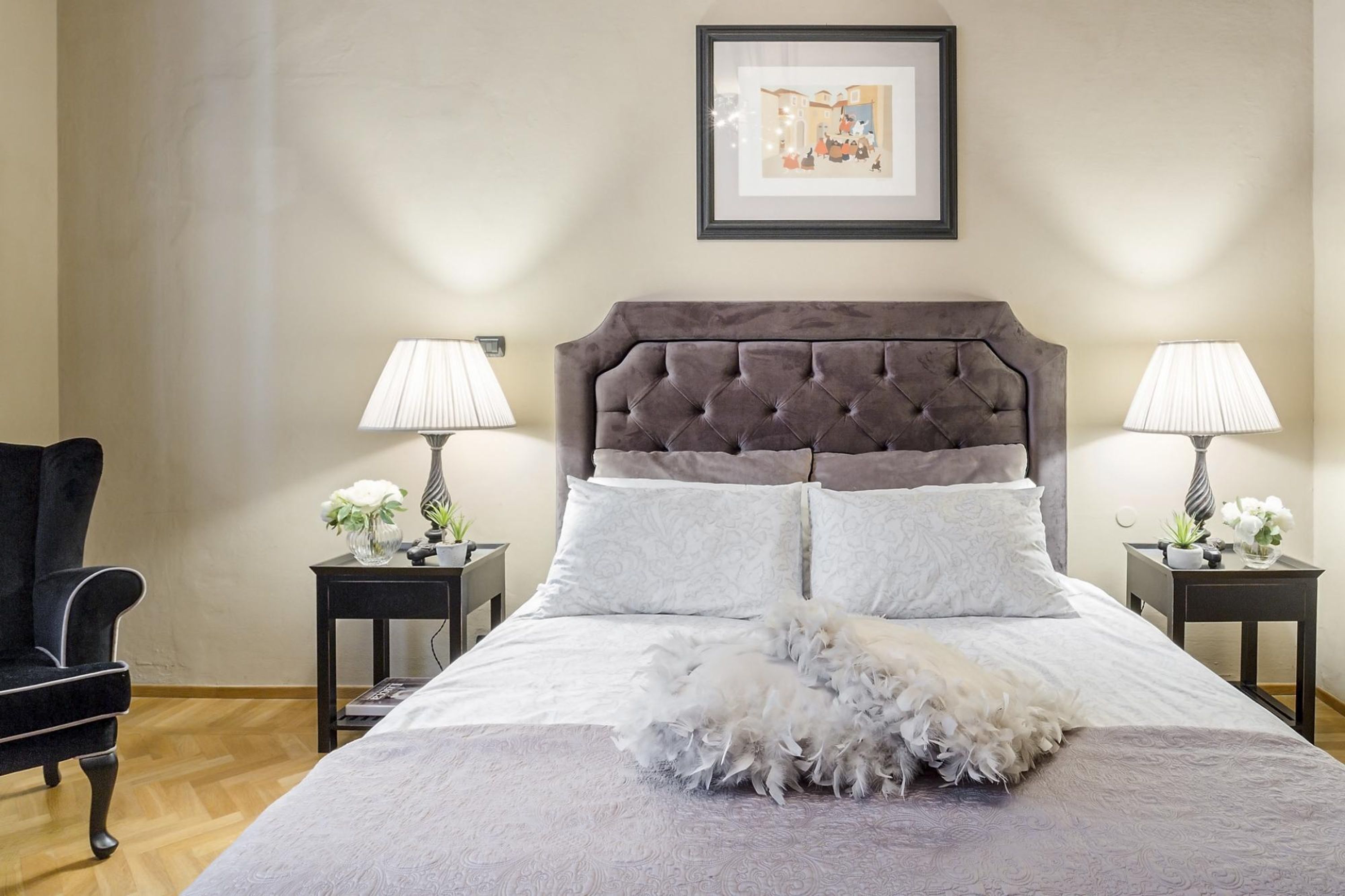 Een netjes opgemaakt slaapkamer met een gestoffeerd hoofdbord, twee nachtkastjes met lampen, een stoel en een ingelijst kunstwerk aan de muur.