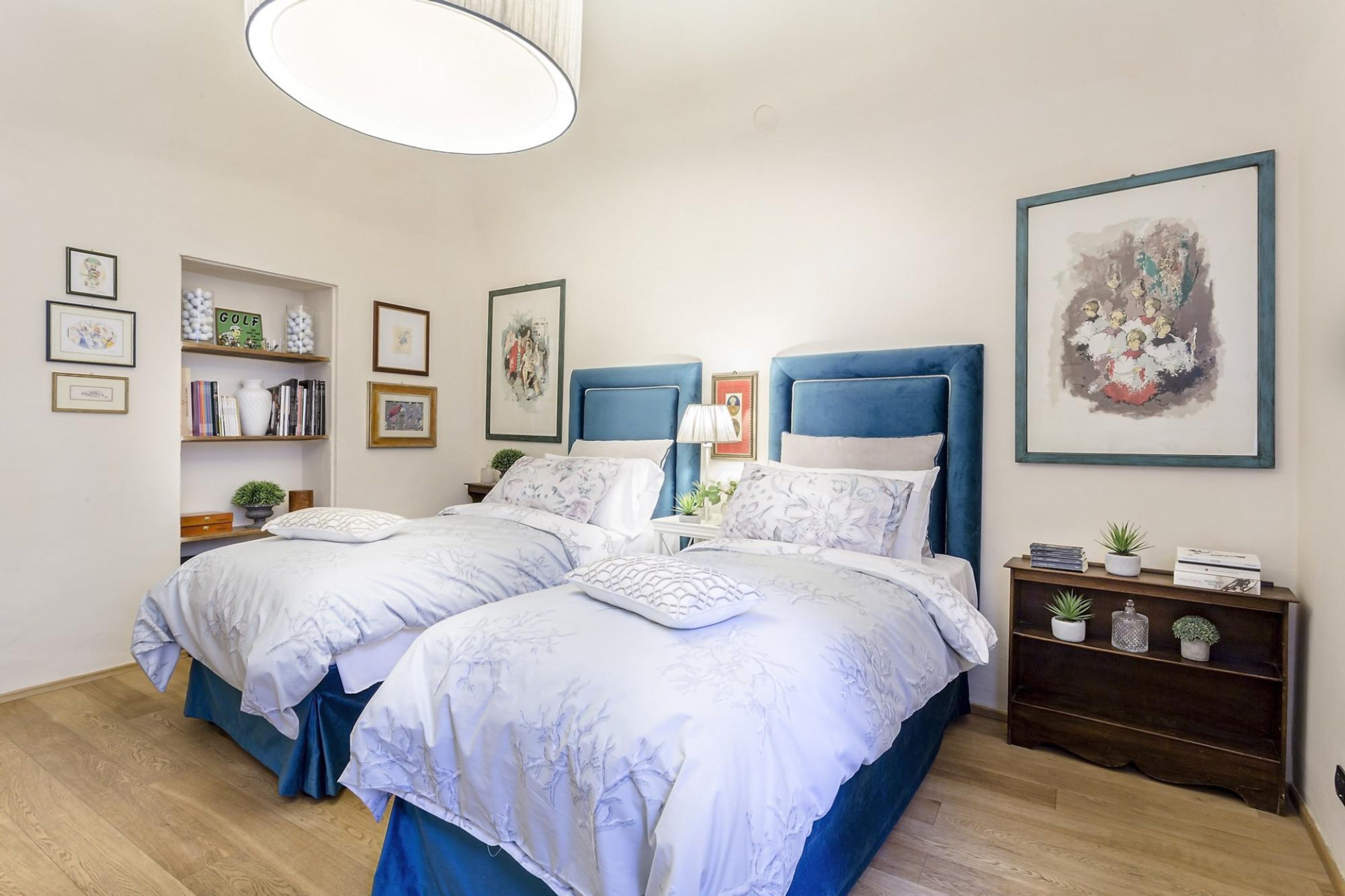 A bedroom with two beds, a bedside table, a lamp, and a shelf with books and decorative items.