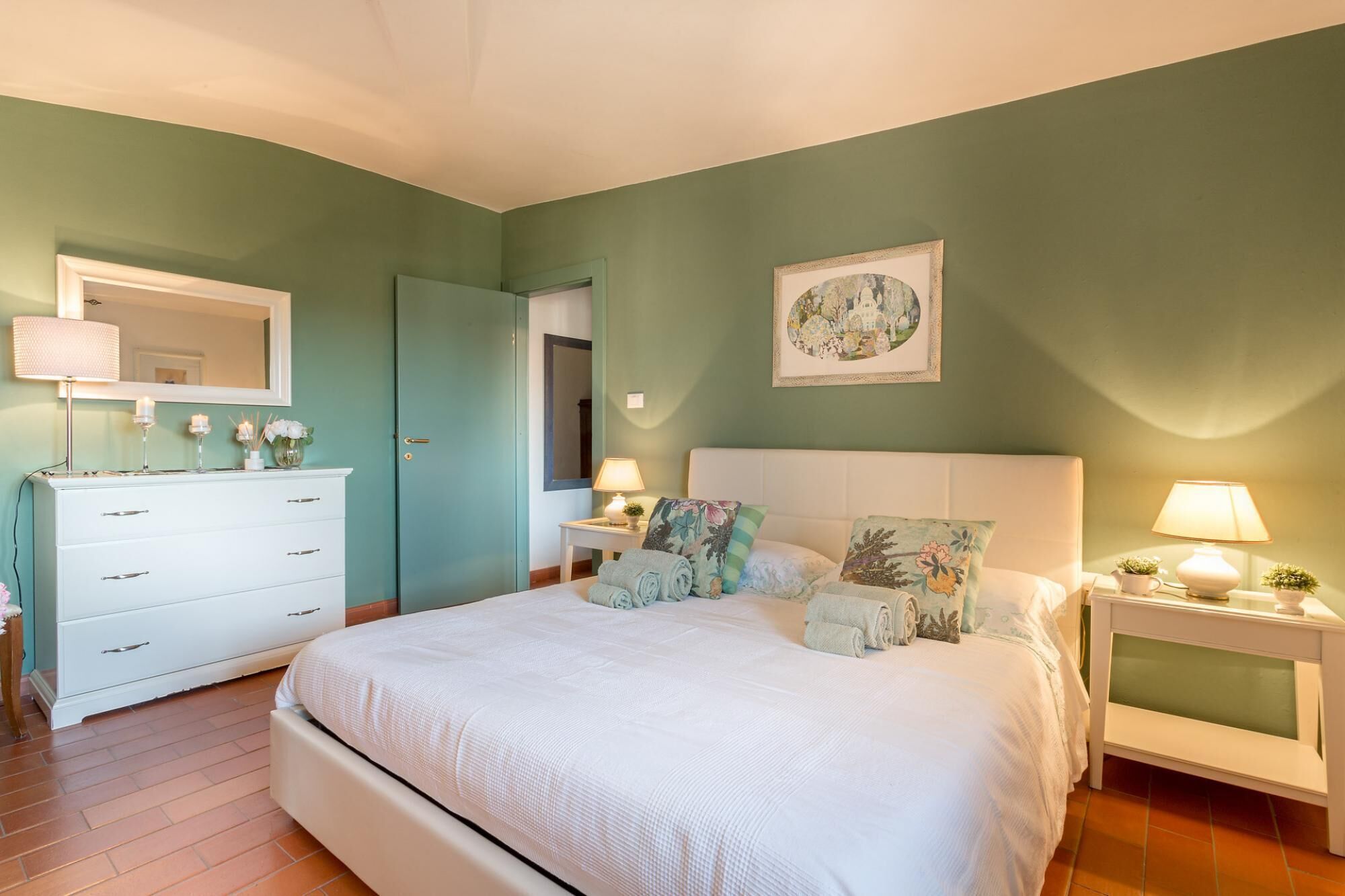 Een slaapkamer met een groot bed, een dressoir, een nachtkastje, een lamp en een lijst met een afbeelding aan de muur.